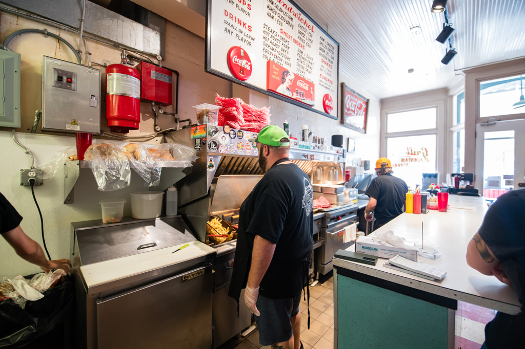 Busy diner kitchen with a cook in a green cap at the grill, menu board and Coca‑Cola signs overhead behind a stainless steel counter