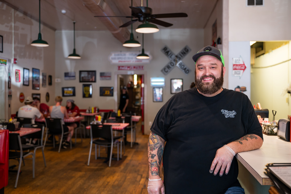 Bearded man in a black t-shirt leaning on a counter inside a retro-style diner, with other diners at red-and-white checkered tables in the background.