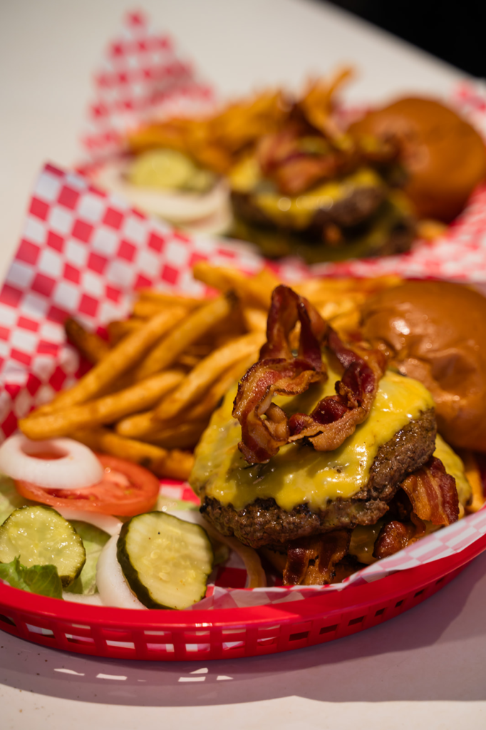 Cheeseburger with crispy bacon on a toasted bun, melted cheese, and a side of fries on a red checkered basket tray with pickles and veggies nearby