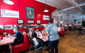 Busy retro diner with red booths, checkered tablecloths, and framed vintage photos; a waitress serves customers at tables.