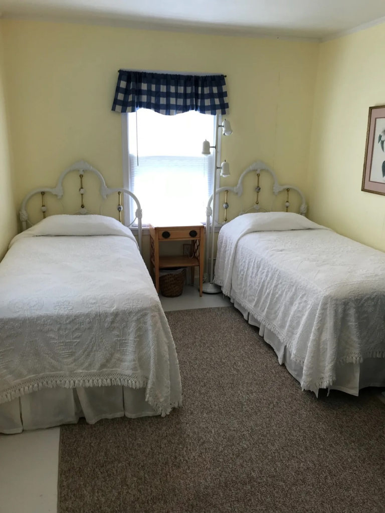 Two white metal-framed twin beds with white bedspreads in a yellow bedroom, a window between them with a blue check valance and a small wooden nightstand between the beds beside a multi-light floor lamp.