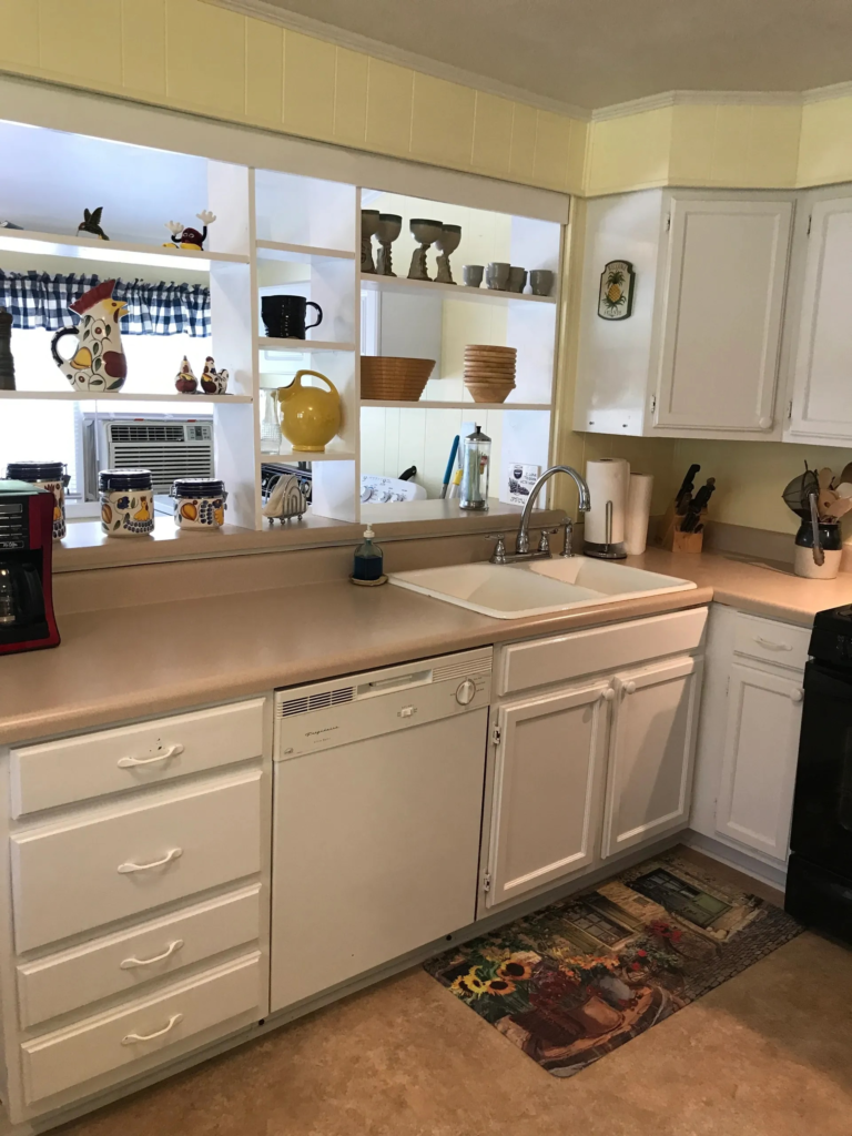 White kitchen with open shelving displaying mugs, bowls, and pitchers above a beige countertop and sink.