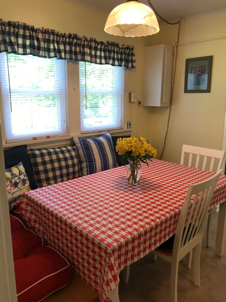 Cozy dining nook with a red-and-white checkered tablecloth, white chairs, and a vase of yellow flowers on the table.
