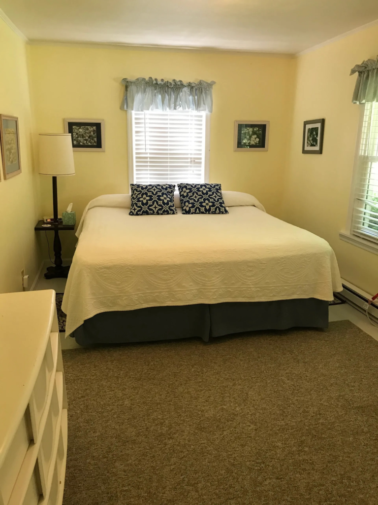 Bedroom with yellow walls and white quilted bed, two navy floral pillows, a lamp on a dark nightstand, and framed wall art by the window with blinds behind the bed