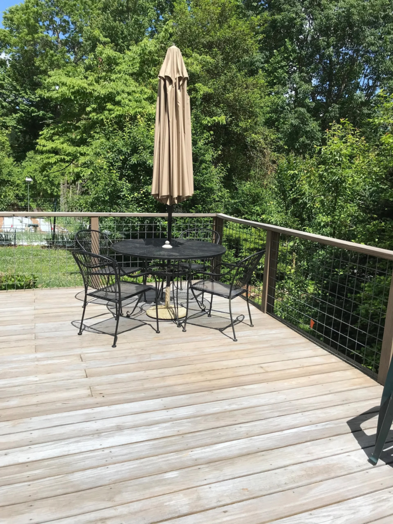 Patio table with four metal chairs and a folded beige umbrella on a wooden deck, surrounded by green trees.