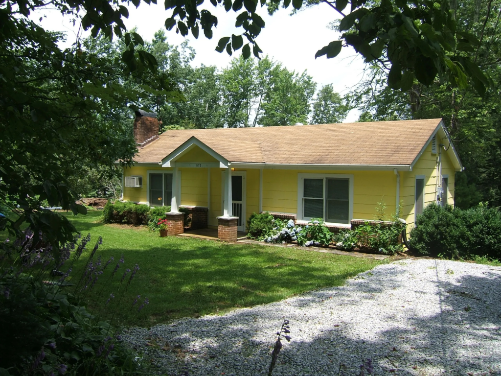 Yellow, single-story house with a brick front porch, surrounded by a green lawn and flowering shrubs under leafy trees.