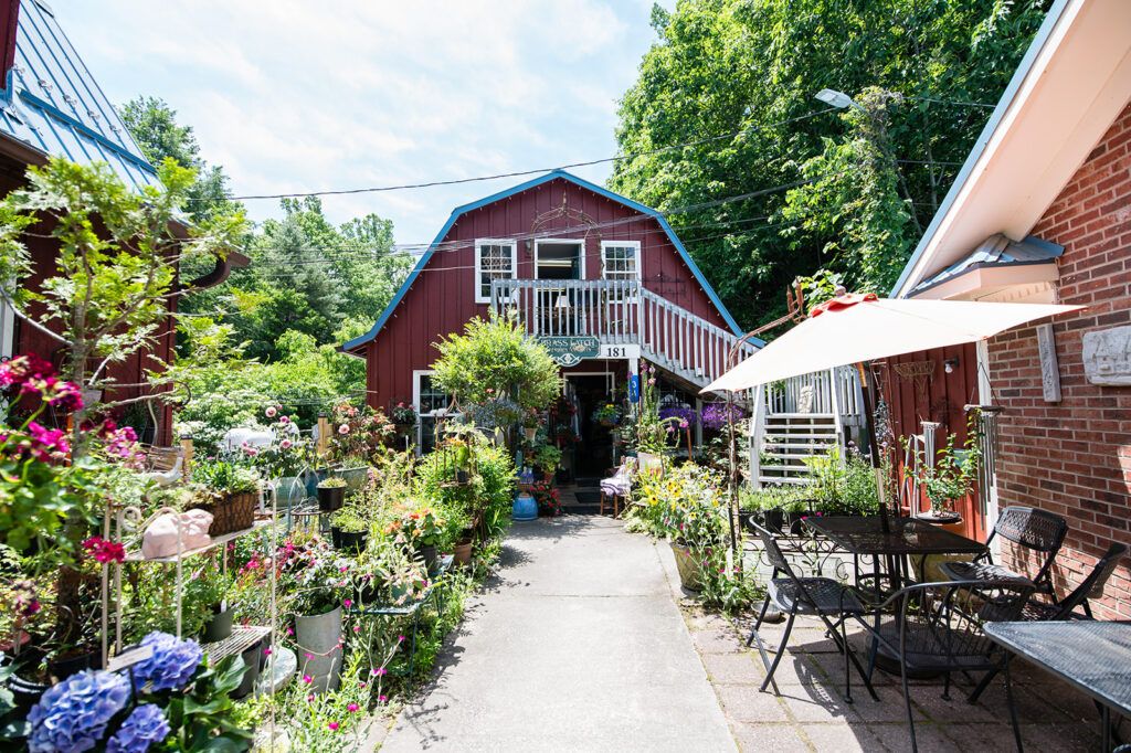 Red barn-style shop with a wooden balcony, surrounded by potted plants and flowers along a paved path. A patio with a table, chairs, and a large white umbrella sits to the right.
