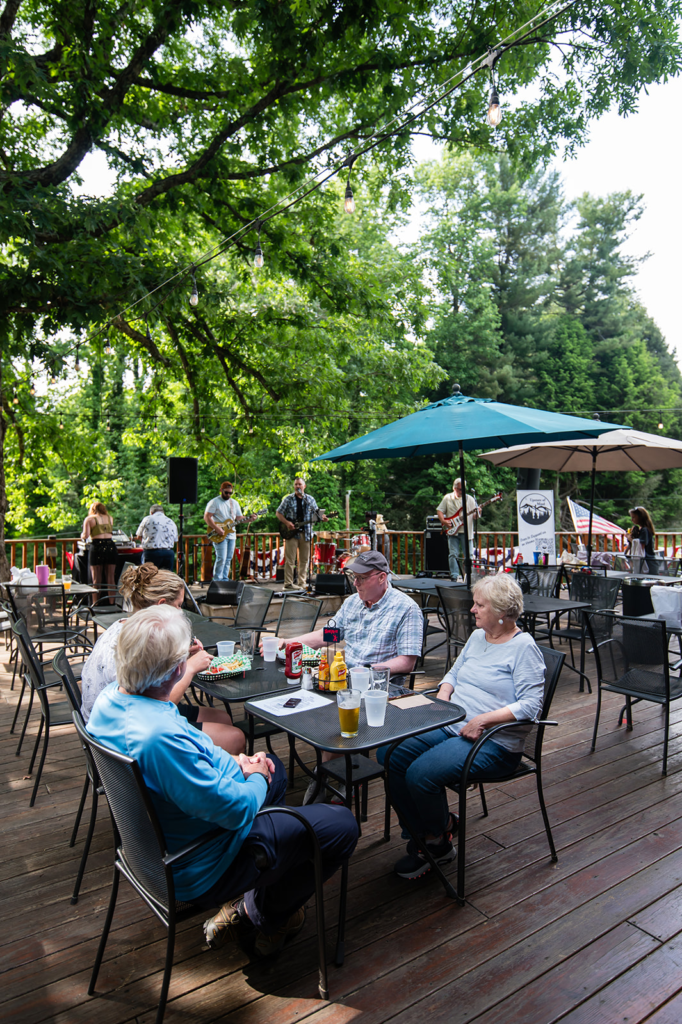 Outdoor wooden patio with a small live band on a stage, string lights overhead, and diners seated at tables under umbrellas by the railing near trees.