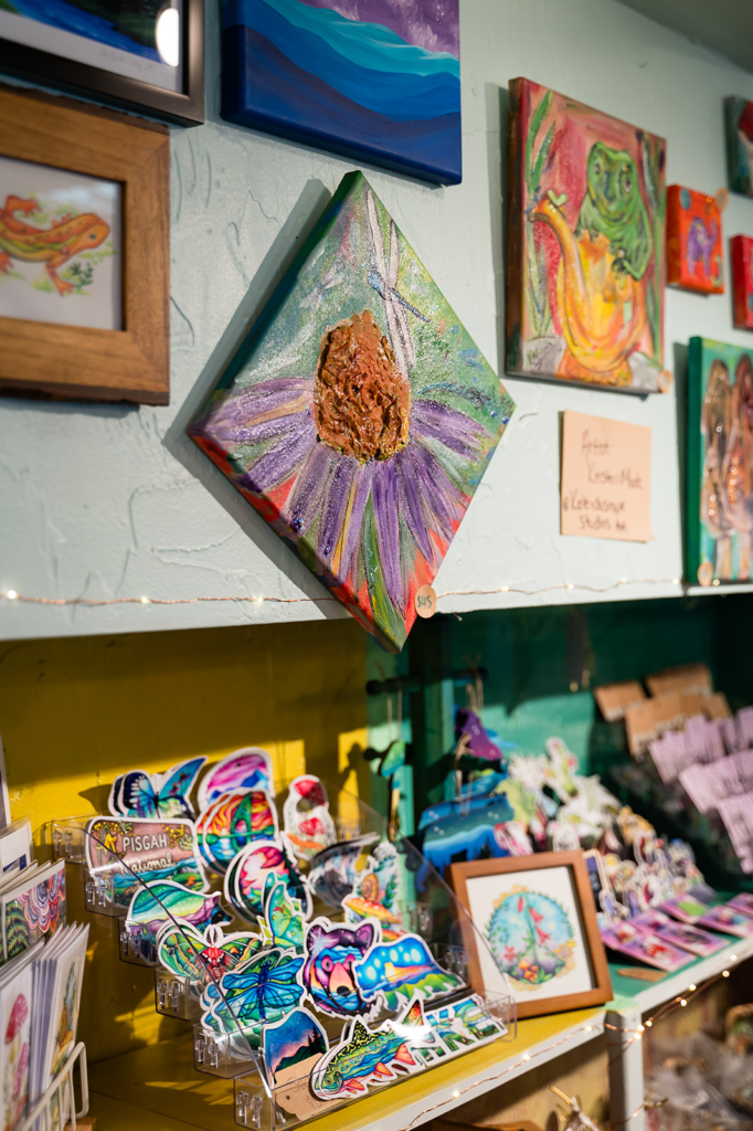 Diamond-shaped painting of a purple flower with a textured brown center and a dragonfly, hung among colorful wall art.
