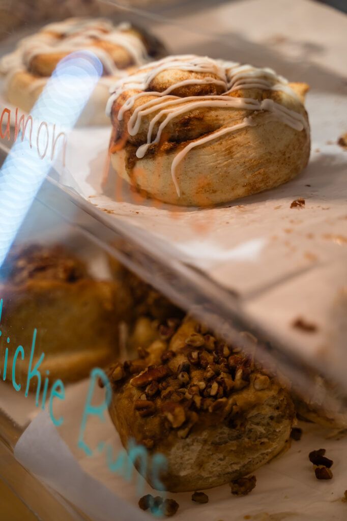 Close-up of a cinnamon roll with white icing in a bakery display case, with another roll and scattered chopped nuts nearby
