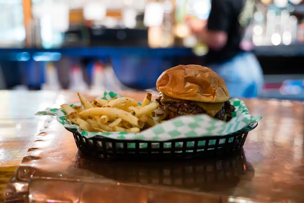 Hamburger with melted cheese and pickles, fries on a checkered paper-lined basket on a wooden counter.