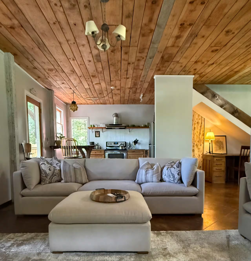 Open-plan living area with a light gray sectional sofa, patterned pillows, and a wood-plank ceiling visible into the kitchen.