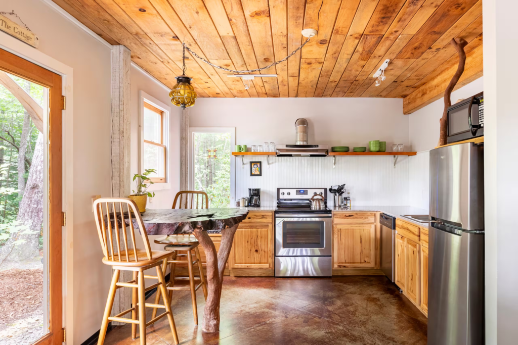 Rustic kitchen with a live-edge wooden island table, wooden chairs, and a wood-paneled ceiling with stainless steel appliances.