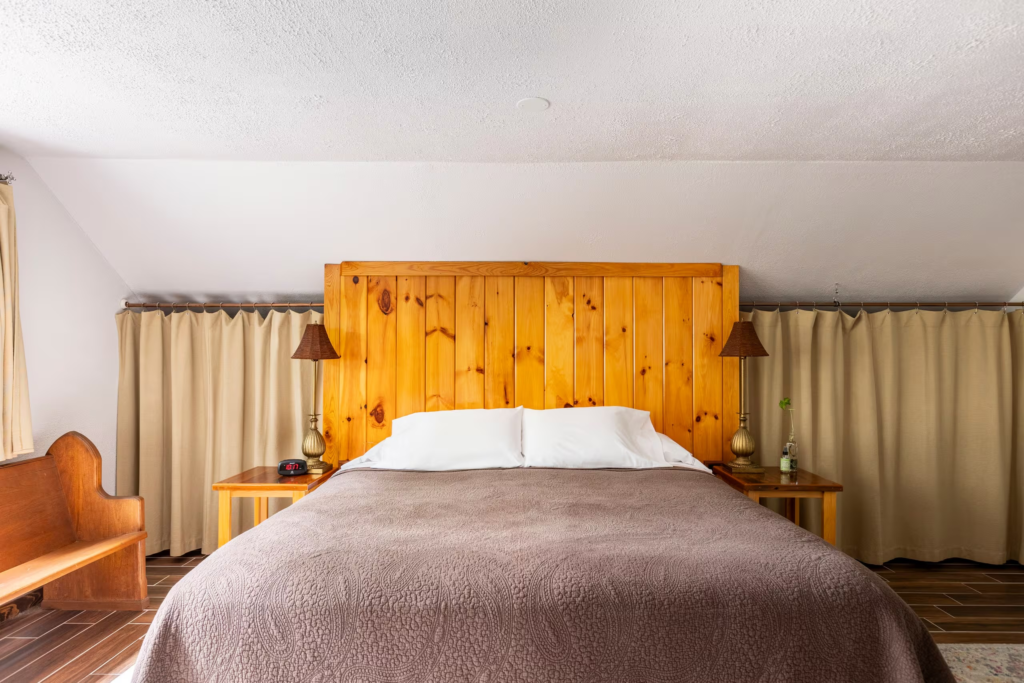 Vacation house bedroom with a tall wooden headboard, two bedside lamps, nightstands, and beige curtains on both sides of the bed.