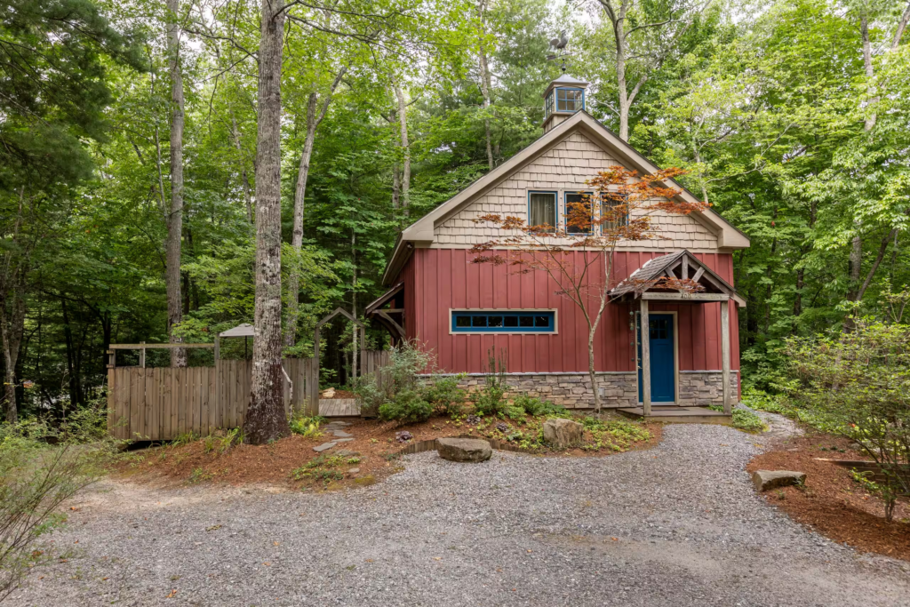 Red wooden cabin nestled in dense green woods with a blue front door and stone foundation.