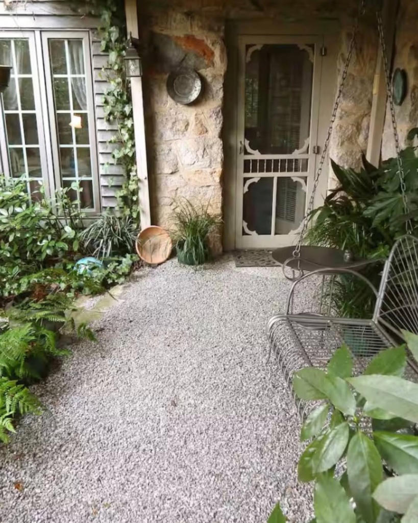 Stone-walled porch with a decorative screened door, gravel path, and a metal swing surrounded by potted plants.