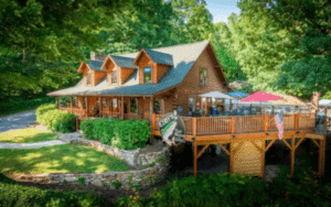 Log cabin-style restaurant on a wooden deck with outdoor seating, green trees, and a pink umbrella.