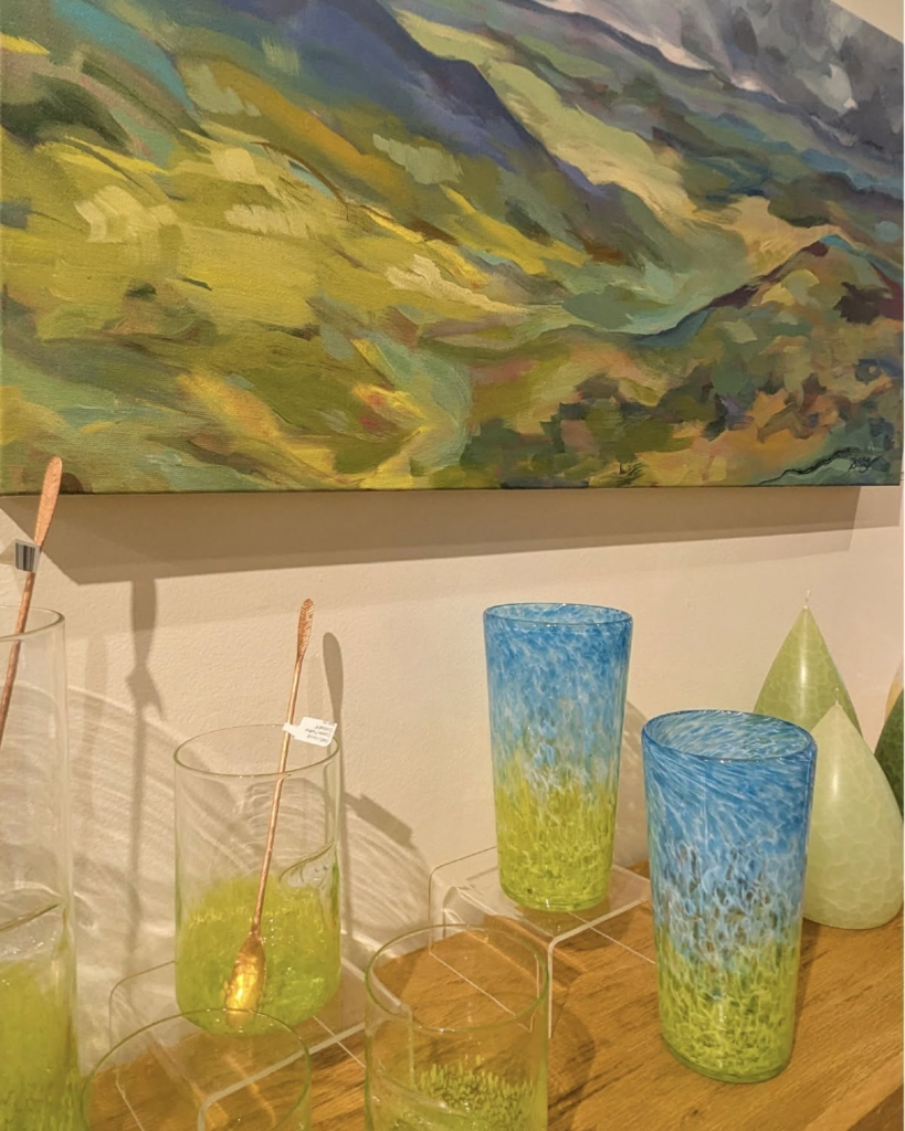Colorful still life display: abstract landscape painting above a collection of gradient glass vases and dishes on a wooden surface.