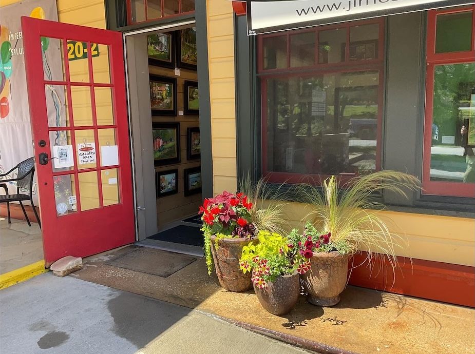 Open red door of a yellow building with flower pots on the doorstep and framed artwork visible inside behind the threshold.