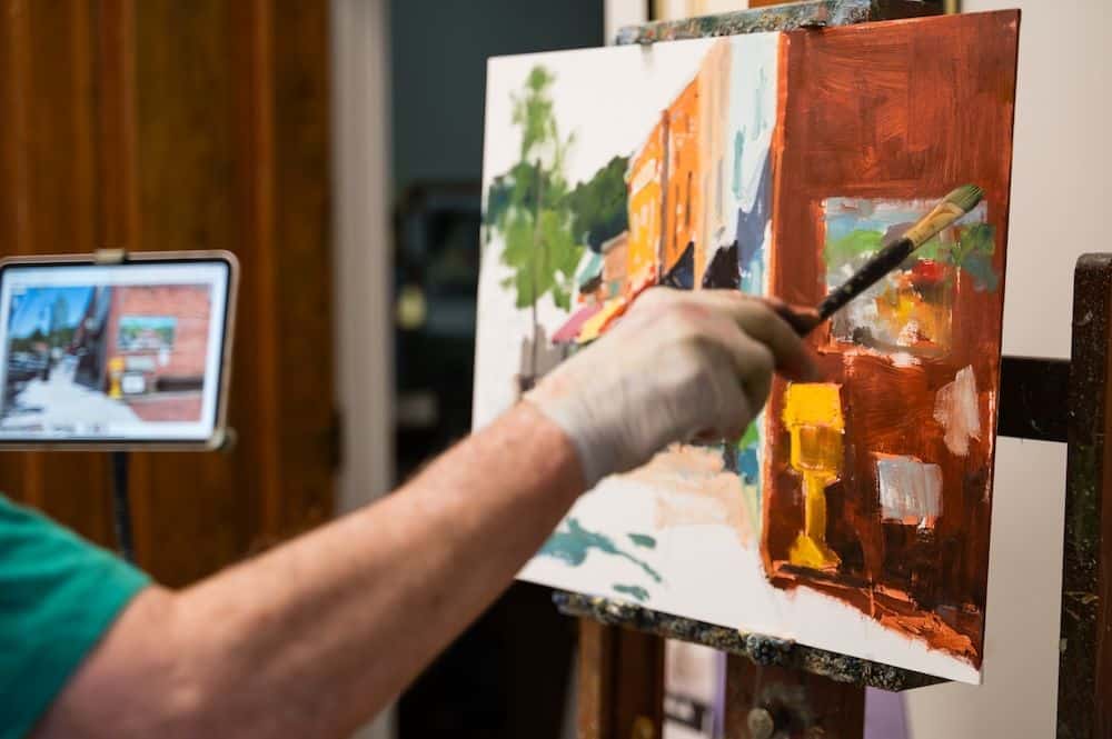 Painter's gloved hand with a brush adding orange paint to a vibrant abstract canvas on an easel in a studio, with a tablet showing a street scene in the background.