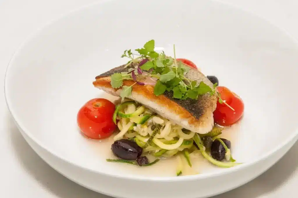 Pan-seared fish fillet with crispy skin resting on a nest of zucchini noodles, cherry tomatoes, olives, and microgreens for garnish