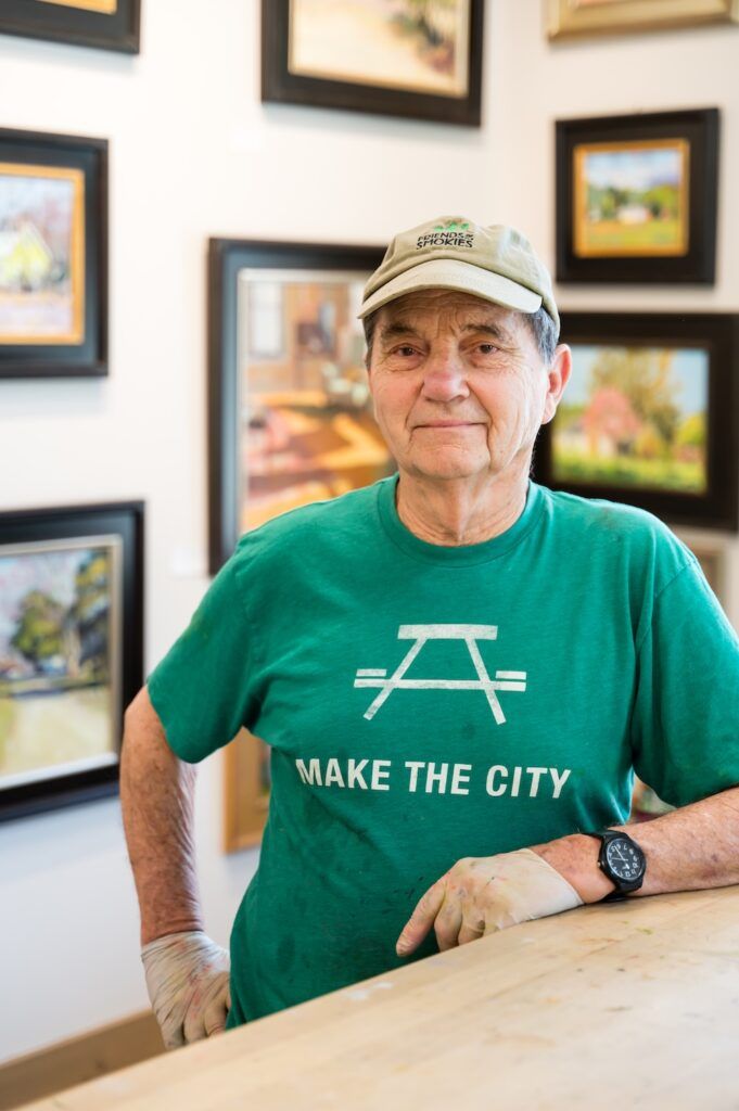 Portrait of an older man in a beige cap and green 'MAKE THE CITY' T‑shirt, standing at a table in an art gallery with framed paintings on the walls.