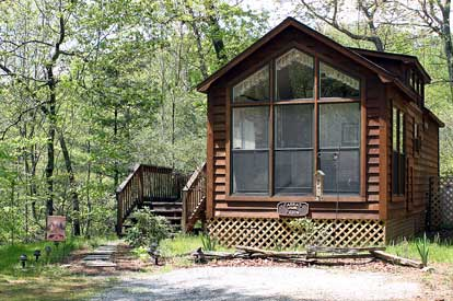 Wooden cabin in a forest setting with large front windows, a triangular peak, and a raised lattice foundation plus a small deck and steps.