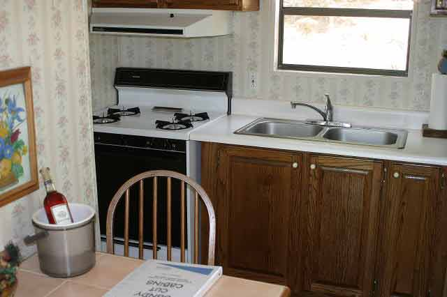 Small kitchen with wooden cabinets, gas stove and range hood, sink, and a window; a table with a chair, a cookbook, and an ice bucket with a bottle.