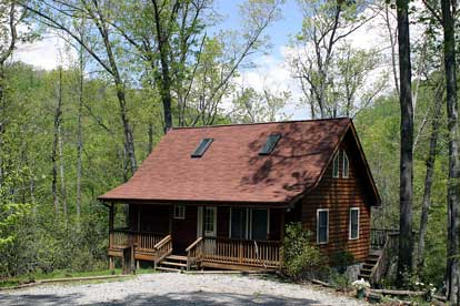 Log cabin with a red roof in a sunlit forest clearing, featuring a front porch and two skylights among tall trees.