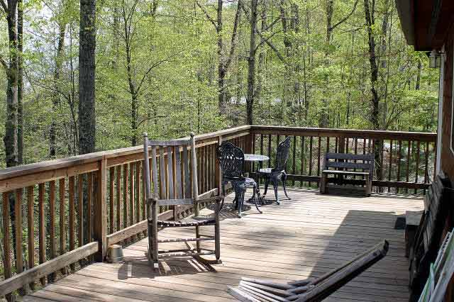 Wooden deck with a rocking chair and a small metal bistro table with two chairs, overlooking a leafy forest.