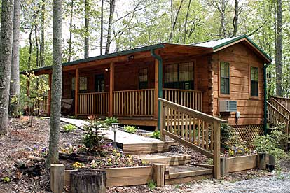 Wooden log cabin with a covered front porch and railings in a wooded yard, featuring a stepped path and raised garden beds along the entry walkway.