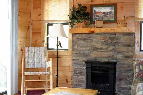 Cozy living room with a stone fireplace, wooden walls, a rocking chair, and a floor lamp by a window.