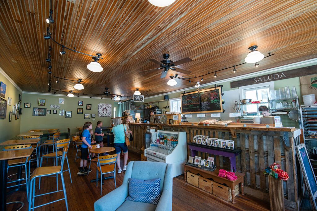 Cozy cafe interior with a wooden ceiling, group of people at the counter near a chalkboard menu and display case of drinks and pastries.