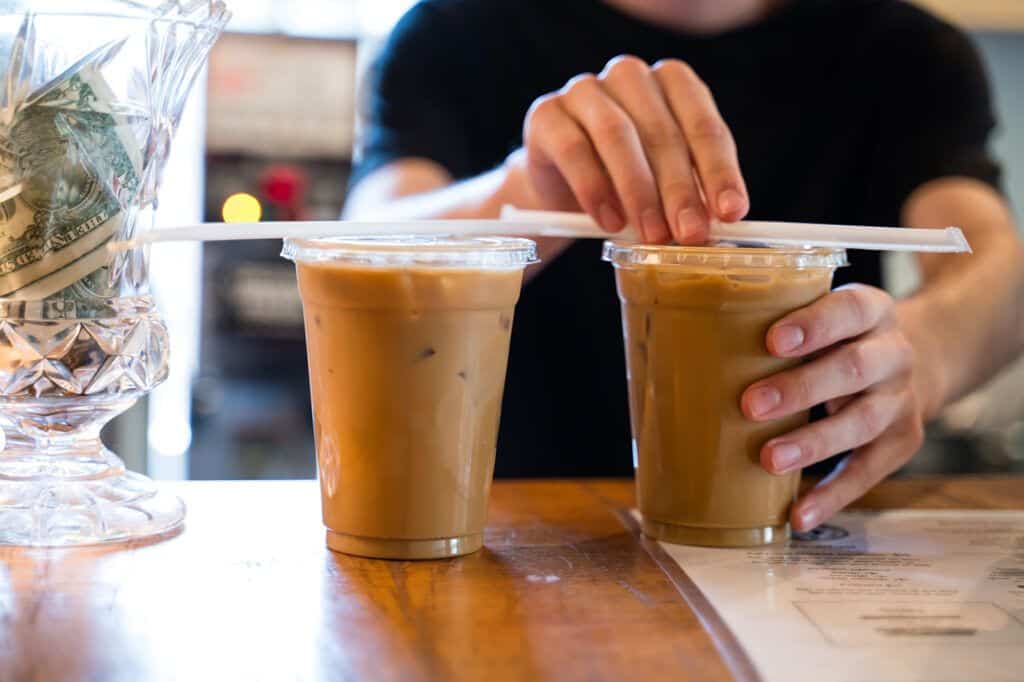 Two iced coffee drinks with plastic lids on a wooden counter, a hand placing a straw between them in a cafe setting.