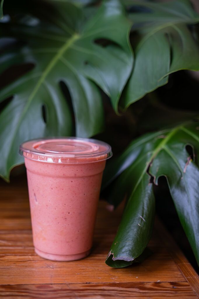 Pink smoothie cup with clear lid on a wooden table, tropical green leaves in the background