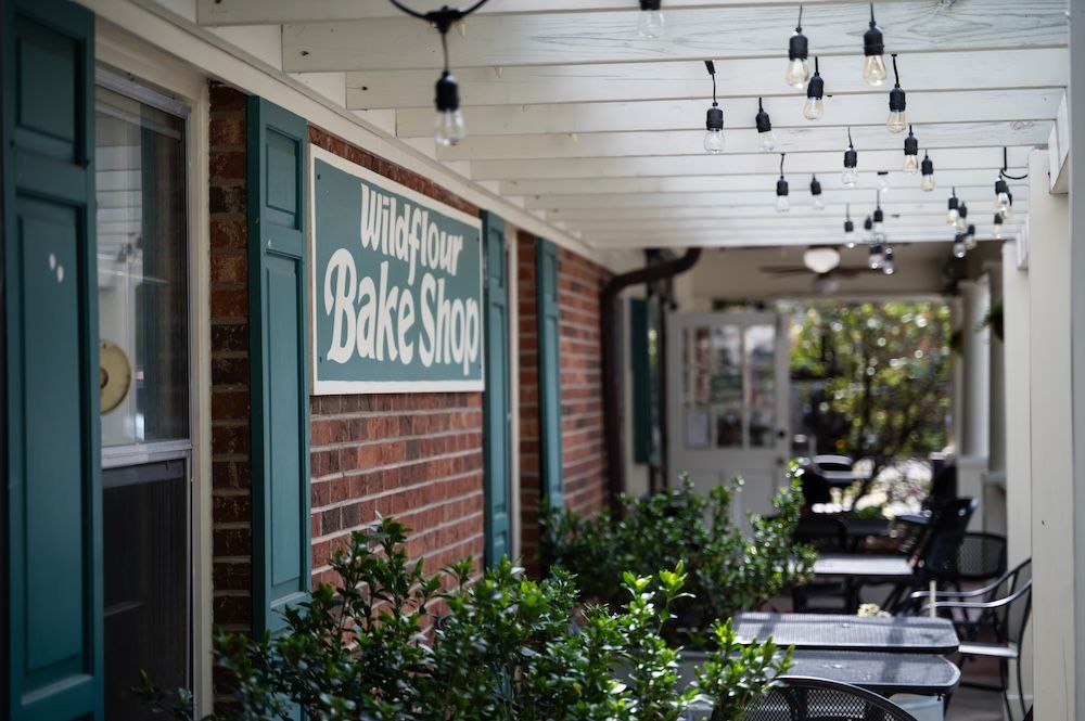 Exterior of Wildflour Bake Shop: brick wall, teal window shutters, and a sign reading 'Wild Flour Bake Shop' under a string of hanging bulbs