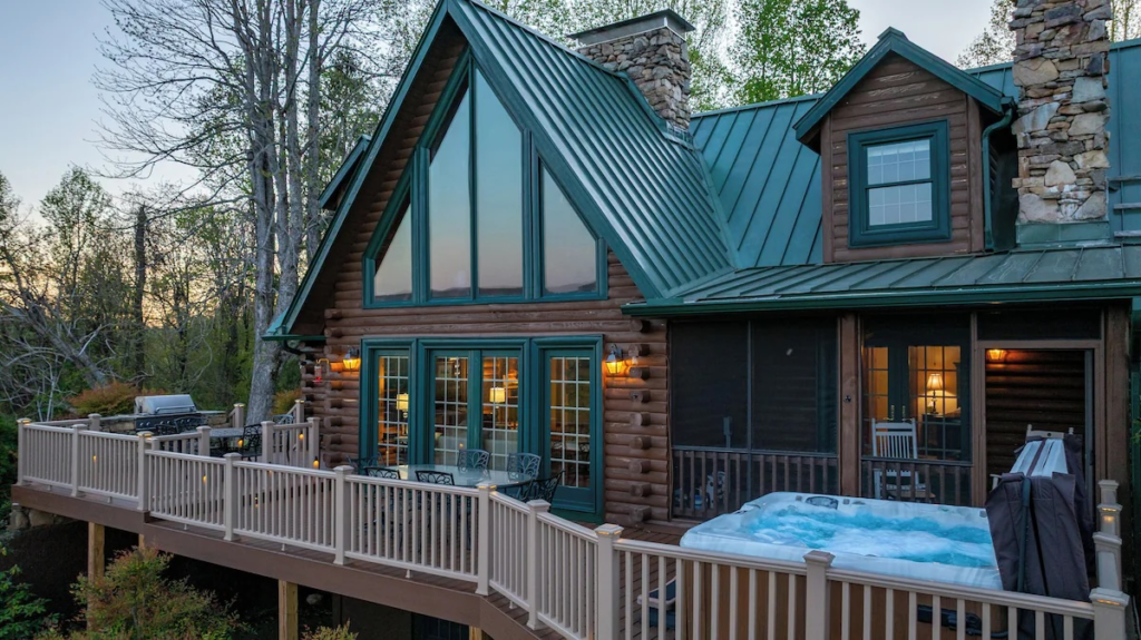Log cabin with teal trim and large glass windows on a wooden deck, hot tub on the railing, surrounded by trees at dusk.