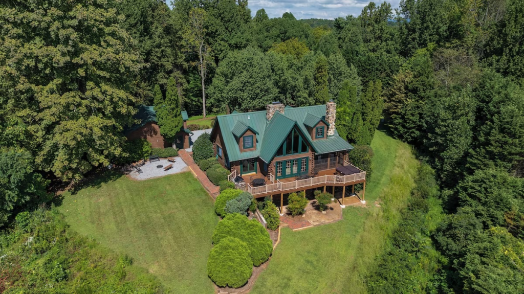 Aerial view of a log cabin–style house with a green metal roof, stone chimneys, and a wraparound deck surrounded by trees.