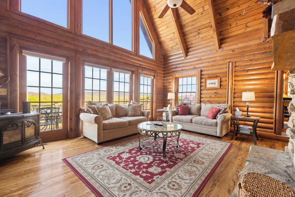 Cozy log cabin living room with beige sofas, a glass-top coffee table, and a red-patterned rug, wood walls and large windows overlooking hills.
