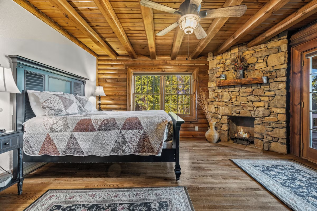 Cozy rustic bedroom with a quilted bed, stone fireplace, and wooden beam ceiling in a log cabin setting.