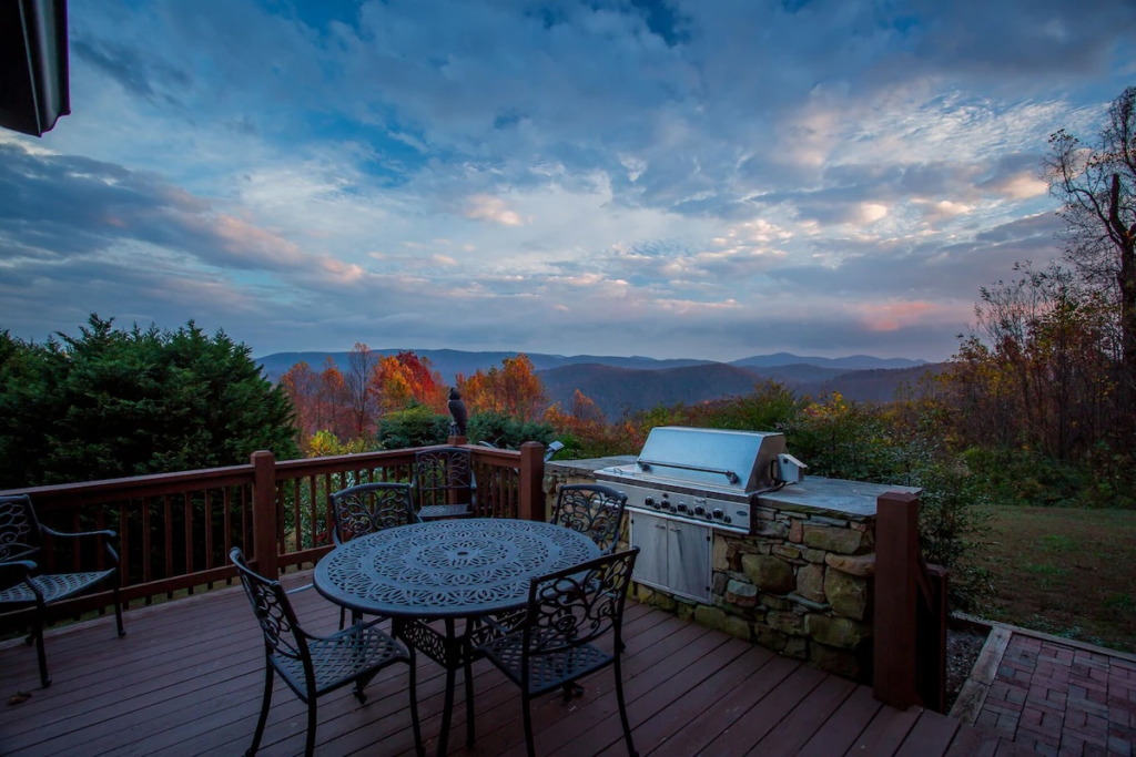 Deck with wrought-iron table and chairs, a stone grill built into a low stone wall, and autumn trees with distant blue mountains under a cloudy sky.