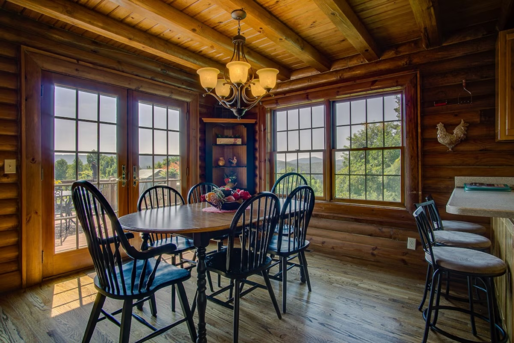 Rustic log cabin dining area with a round table and black windsor chairs by large windows and french doors, wood floors, and a chandelier.
