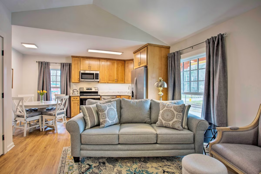 Open-concept living room with light gray sofa, patterned pillows, and a front-facing view of a wood kitchen and dining area.