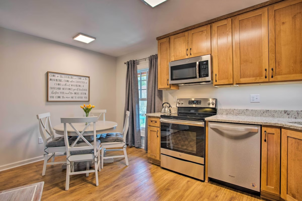 Cozy kitchen with oak cabinets, stainless steel stove and dishwasher, and a white round dining table with four chairs and a yellow tulip centerpiece.