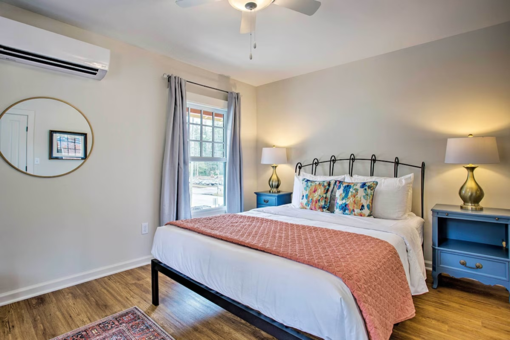 Queen bed with white bedding, peach quilt, and colorful pillows flanked by blue nightstands with brass lamps on either side.