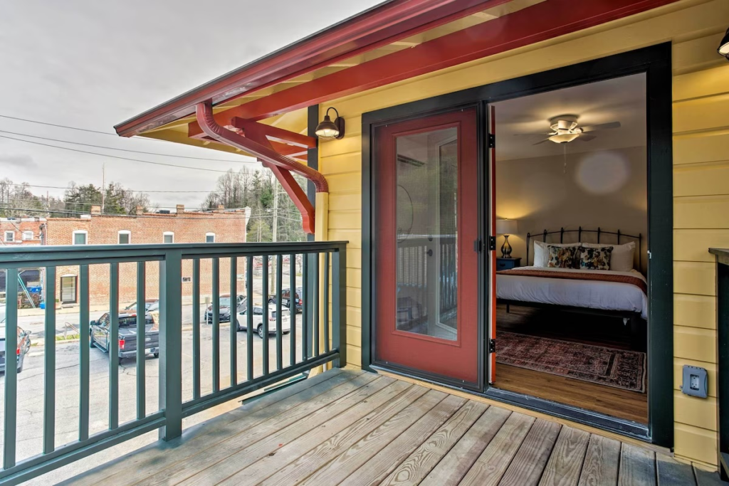 Open red-framed door leading to a cozy bedroom with a bed, rug, and ceiling fan seen from a yellow building balcony exterior.