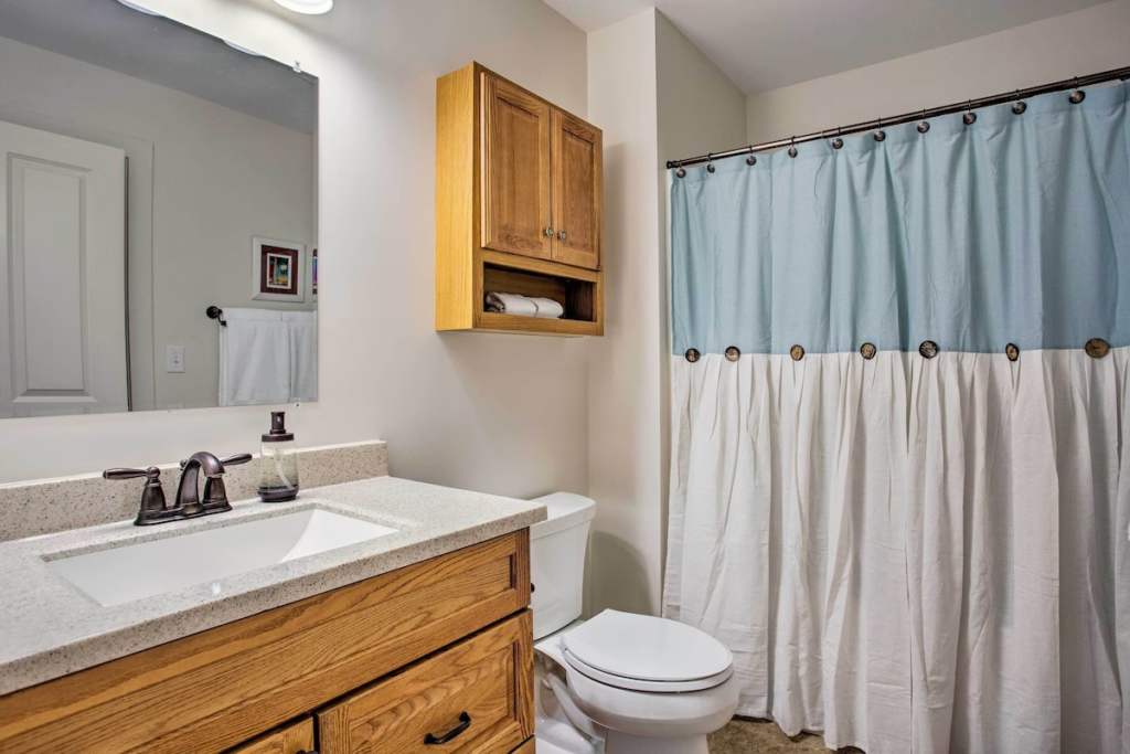 Bathroom with a wood vanity, sink, and beige countertop, soap dispenser nearby, mirror overhead, and a toilet beside a blue-and-white shower curtain.