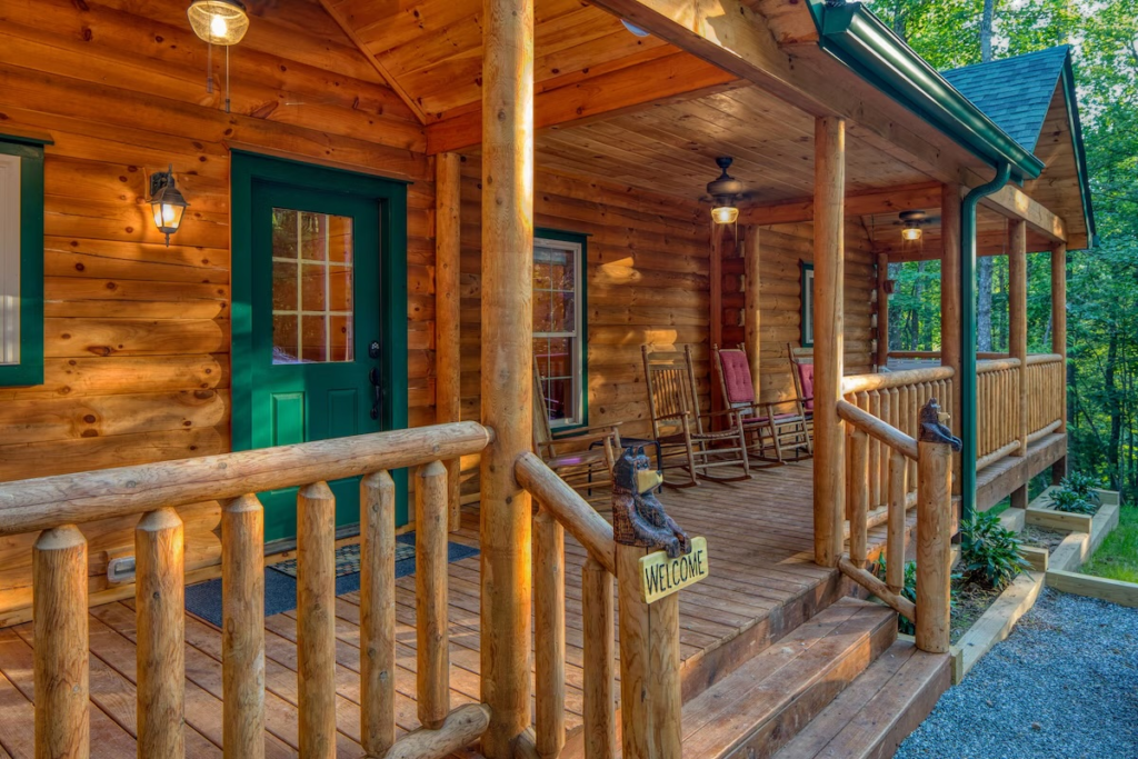 Front porch of a log cabin with a green door, wooden railings, and rocking chairs under a covered porch.