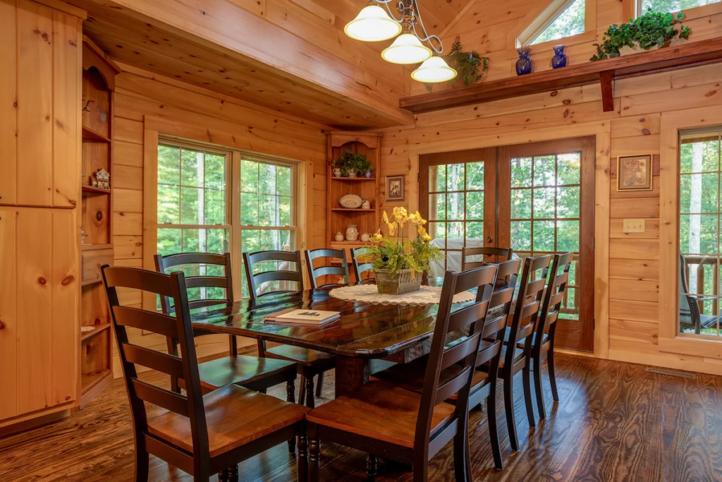 Rustic cabin dining room with a long dark wooden table and multiple chairs, wood-paneled walls, large windows, and french doors showing greenery outside.