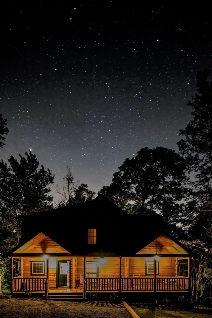Night scene of a wooden house with warm porch lights beneath a clear, star-filled sky surrounded by trees.